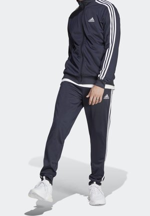 Mężczyzna ubrany w czarny dres Adidas z białymi paskami i logo, w połączeniu z białymi sneakersami, stoi na białym tle.