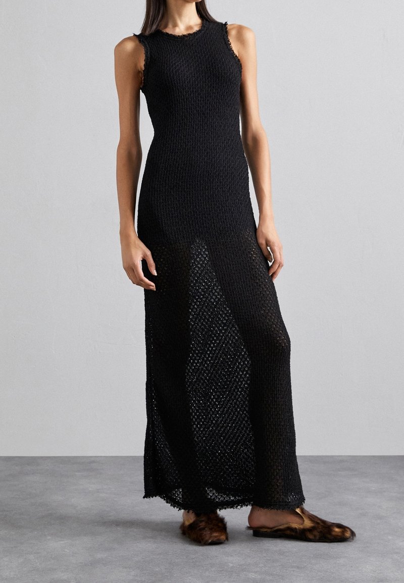 Robe maxi noire tricotée sans manches, avec une texture en maille ouverte et des bords frangés. Associée à des chaussures slip-on en fourrure.