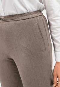 Braune Stretchhose mit taillierter Passform, elastischem Bund, Seitentaschen und glattem Textil. Kombiniert mit einem weißen Hemd.