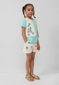 Hellblaues T-Shirt, cremefarbene bestickte Weste mit Blumen, hellgelbe Shorts mit verschiedenen bunten Mustern und goldene Sandalen mit Riemchen.