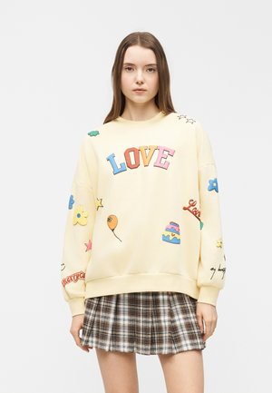 Jeune femme portant un sweat-shirt crème avec un texte coloré "LOVE" et des patchs ludiques, associé à une jupe plissée à carreaux marron et blanc.
