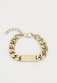 Bracelet en métal doré avec des maillons en chaîne entrelacés, présentant une plaquette rectangulaire gravée du logo Lacoste et un fermoir.