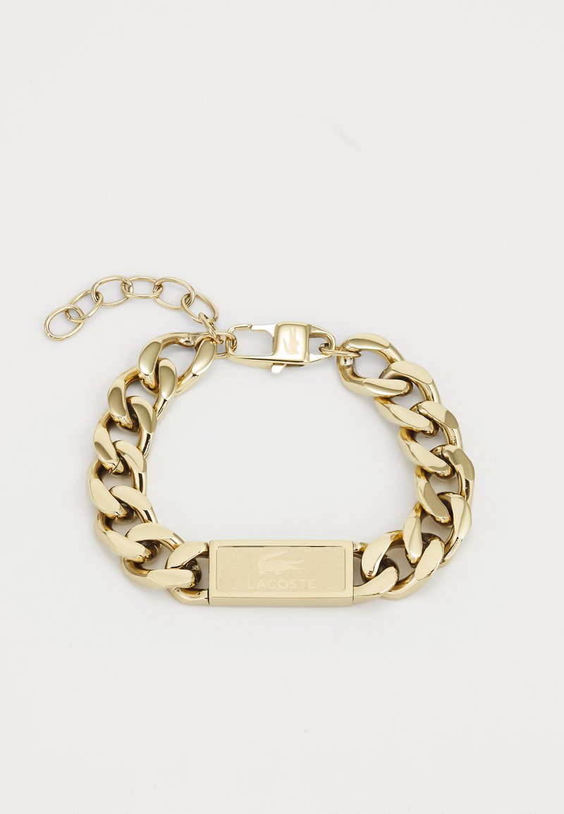 Bracelet en métal doré avec des maillons en chaîne entrelacés, présentant une plaquette rectangulaire gravée du logo Lacoste et un fermoir.