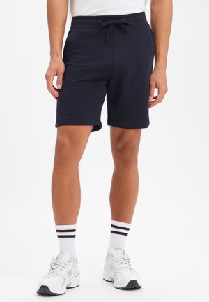 Mann, der schwarze Shorts mit Kordelzug, weiße Sportsocken mit schwarzen Streifen und weiße Turnschuhe trägt, steht vor einem schlichten Hintergrund.