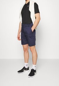 Černé polo tričko, tmavě modré šortky, bílý svetr přehozený přes ramena a černé tenisky s bílou podrážkou. Na tričku a botách je značka Nike.