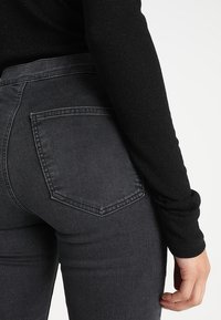 Osoba oblečená v tmavě šedých džínech s vysokým pasem a dlouhém černém topu, s jednou rukou uvolněně po boku.