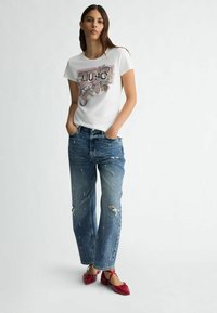 T-shirt bianco in cotone con design grafico colorato e ornamentale; jeans blu stracciati con decorazioni; scarpe rosse con punta affusolata.