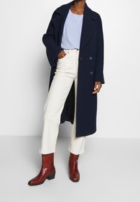 Cappotto doppiopetto in misto lana blu navy, jeans a gamba larga color crema e stivaletti in pelle bordeaux con tacco blocco. Texture morbide e vestibilità su misura.