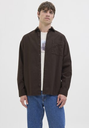 Hombre con camisa marrón oscuro desabrochada sobre camiseta gráfica blanca y vaqueros azules, de pie contra un fondo claro y liso.