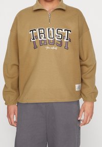 Khakifarvet pullover med lynlåshals, med teksten "TRUST" i fede bogstaver og en mindre tekst nedenunder. Ribbet tekstur og elastiske manchetter.