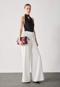 Top noir sans manches avec un décolleté drapé, associé à un pantalon large blanc. Le modèle tient un sac à main en cuir bordeaux, debout sur un sol gris.