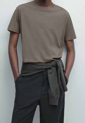 Personne portant une chemise marron à manches courtes, un pantalon noir, avec un pull gris foncé noué autour de la taille, les mains dans les poches.