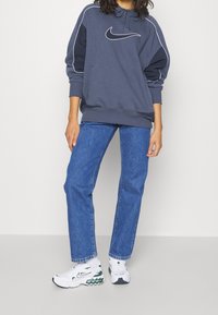 Navy blue hoodie with white stitching and a front pocket, paired with high-waisted blue jeans and white sneakers featuring green accents.