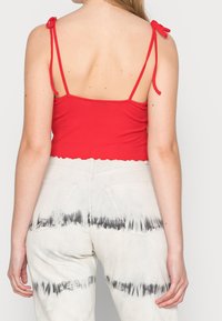 Červený žebrovaný crop top s tenkými ramínky a zoubkovaným lemem, nošený s světle šedými high-waisted džínami s tie-dye vzorem a zadními kapsami.