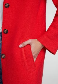 Mano infilata nella tasca anteriore di un cappotto rosso brillante con bottoni, indossato sopra jeans e una maglietta bianca.