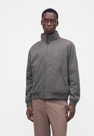 Man wearing a gray zip-up jacket with pockets and brown pants, standing against a plain white background.