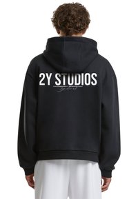 Juoda su kapuce megztinis, kuriame nugaros pusėje yra didelis baltas grafiškas tekstas "2Y STUDIOS", su laisvu kirpimu ir plačiomis rankovėmis.