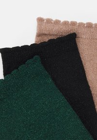 Tre par glitrende ribbede sokker i mørkegrøn, sort og lysebrun med bølgede kanter. Hver sok har en struktureret finish.