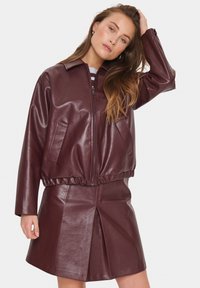 Giovane donna con lunghi capelli, che indossa una giacca di pelle borgogna con zip e una gonna di pelle abbinata, mentre posa con una mano sulla testa.