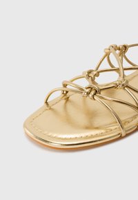 Gold metallic sandal featuring a flat design with intricate crisscross straps, a rounded toe, and stitched detailing along the edge.