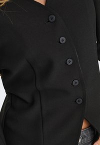 Vêtement noir asymétrique à boutons porté sur un jean, montrant la couture latérale et cinq boutons ronds noirs visibles.