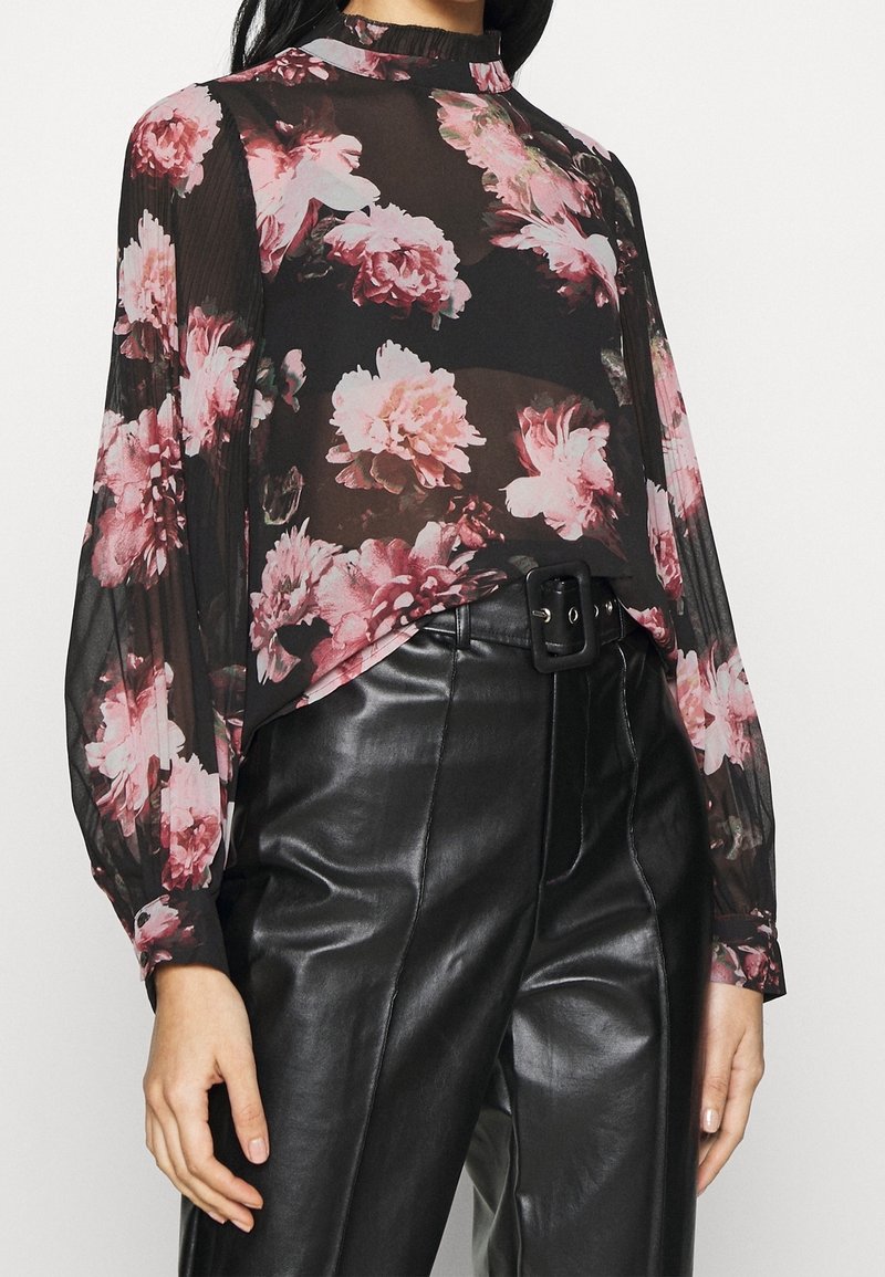 Femme portant un chemisier noir transparent à motifs floraux avec manches longues, rentré dans un pantalon en cuir noir taille haute avec une grande boucle de ceinture.