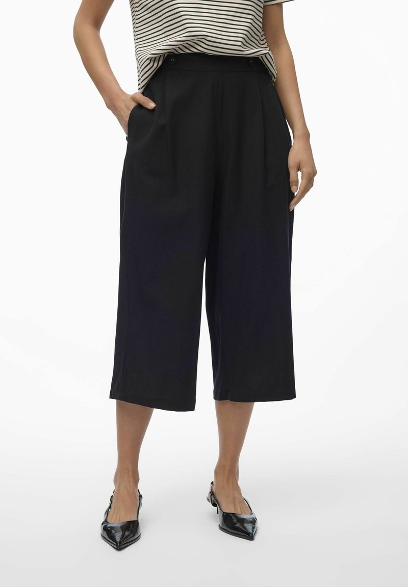 Femme portant un pantalon noir large coupé au-dessus de la cheville, un haut à manches courtes rayé, et des chaussures slingback noires à bout pointu, mains dans les poches.