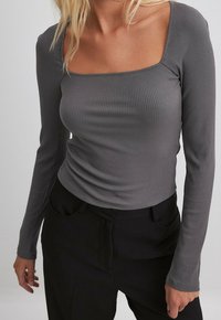 Femme portant un haut gris côtelé à manches longues ajusté avec un décolleté carré et un pantalon noir, debout devant un fond uni.