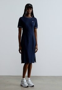 Tumši zila kokvilnas kleita ar īsām piedurknēm, kontrastējošu apdari un sānu šķēlumiem. Valkāta kopā ar melnu cepuri, baltām potīšu zeķēm un masīvām sporta kurpēm.