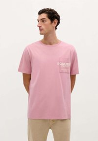 Růžové bavlněné tričko s kulatým výstřihem, krátkými rukávy a kapsičkou na prsou se bílým textem. Jednoduchý design s hladkou texturou.
