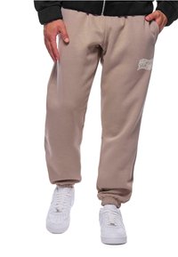 Beige sweatpants med elastiske manchetter, sidelommer og en trykt logo-detalje nær hoften, parret med hvide sneakers.