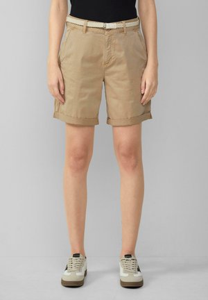 Shorts en coton beige avec des ourlets retroussés, dotés de poches latérales et d'une ceinture tressée blanche. Associés à des sneakers de couleur claire.