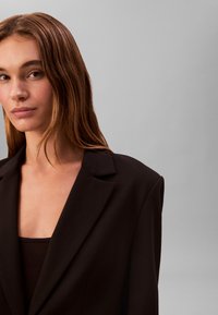 Sort struktureret blazer med skarpe revers og skræddersyet pasform, kombineret med en sort top, sat mod en neutral baggrund.