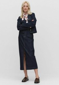 Blondi nainen seisoo kädet ristissä, päällään tumma farkkutakki, sen kanssa matching pitkä hame jossa on etureikä, valkoinen paita, kuvioitu kaulahuivi ja ruskeat loaferit.