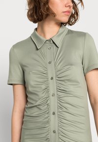 Blusa verde-claro de manga curta com gola e fecho dianteiro por botões, apresentando detalhe de tecido franzido ao longo do corpete.