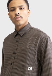 Brown checkered button-up shirt with a collared neck, a single chest pocket, and black buttons; features a logo tag on the pocket.