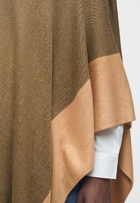 Poncho tissé marron et beige drapé sur la manche blanche d'une chemise et la main à la peau foncée d'une personne.