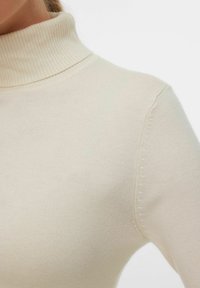 Jersey de cuello alto color crema con cuello acanalado, textura de tejido suave y detalles sin costuras en los hombros.