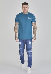 Teal cotton t-shirt with a small white logo on the chest, paired with distressed blue jeans featuring patchwork and rips, and white sneakers.