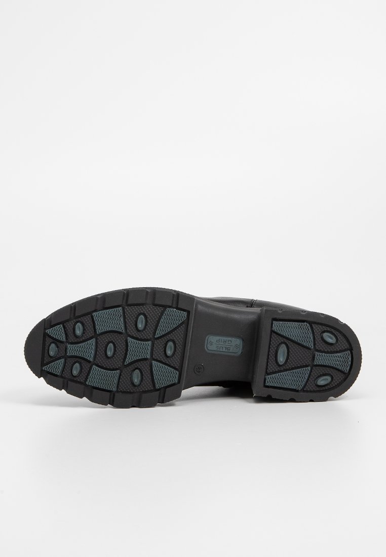 Suola nera di una scarpa con grip in gomma a motivo, caratterizzata da scanalature ovali e rettangolari per la trazione, fotografata su uno sfondo bianco.