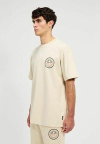 Camiseta de algodón beige con mangas cortas, cuello redondo y parche con logo redondo en el pecho que presenta un diseño gráfico. Corte recto y ajuste casual.