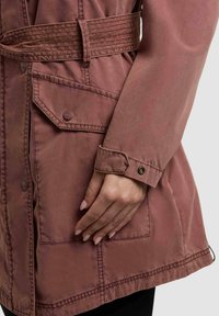 Približni posnetek rokava prašno rožnate jakne z manšetnim zapenjanjem na sponko in roko, ki počiva blizu žepka z zaponko na sponko.