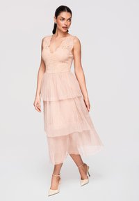 Femme en robe midi en dentelle et tulle rose poudré superposée avec décolleté en V, posant en talons crème à brides à la cheville sur fond uni.