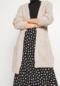 Beżowy dzianinowy kardigan z kapturem, otwarty z przodu, z prążkowanymi mankietami i bocznymi kieszeniami, noszony na czarnej sukience w kremowo-brązowy wzór w groszki.