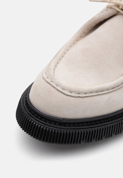 Beige Wildlederschuh mit runder Zehenpartie, schwarzer Gummisohle mit gerippter Textur und kontrastierenden Nähten an den Kanten.