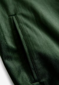 Tessuto verde scuro con una texture liscia; presenta una tasca laterale discreta con cuciture pulite, che mette in risalto un design raffinato e snodato.