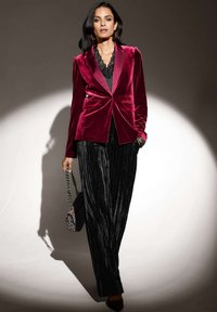 Bordeaux fluwelen blazer met piek-lapels, gecombineerd met zwarte, geplooide, wijde broeken. Geaccentueerd met een kleine tas en textuurhaar.