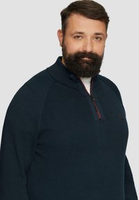 Marineblauwe pullover trui met een gestructureerde brei, hoge kraag en een kwart-rits aan de voorkant. Voorzien van een contrasterende rode rits en logo-accent.