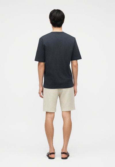 Άνδρας που φοράει απλό μαύρο κοντομάνικο μπλουζάκι, μπεζ σορτς μέχρι το γόνατο και μαύρα σανδάλια, στέκεται με την πλάτη προς τα εμάς σε λευκό φόντο.