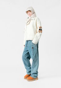 Chaqueta de mezcla de lana blanca con capucha y parche de oso de peluche, pantalones de mezclilla azul de corte holgado y calzado de ante marrón. Acentos en gris claro en la capucha.